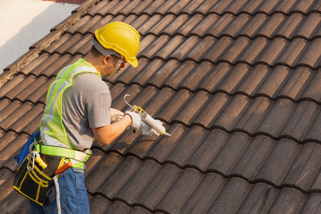 A worker in a hard hat and safety vest seals gaps between brown roof tiles with a caulk gun.