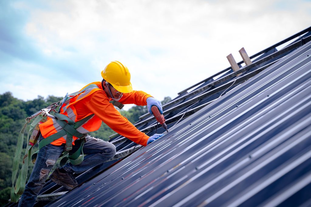 A construction worker in safety gear installs solar panels on a dark roof with a cloudy sky behind.
